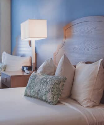 Two neatly made twin beds with wooden headboards, white pillows, and decorative green throw pillows beside a nightstand with a lamp and phone, against a blue wall and window with white shutters.