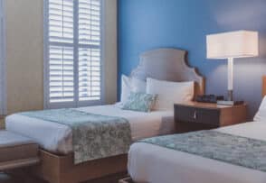 A hotel room with two single beds, green floral bed runners, a wooden nightstand with a lamp, and white shuttered windows.