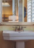 A bathroom sink with a bronze faucet is positioned below a large mirror framed in gold. There is a toilet paper holder on the left wall and white shutters on the right window.