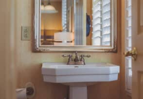 A bathroom sink with a bronze faucet is positioned below a large mirror framed in gold. There is a toilet paper holder on the left wall and white shutters on the right window.