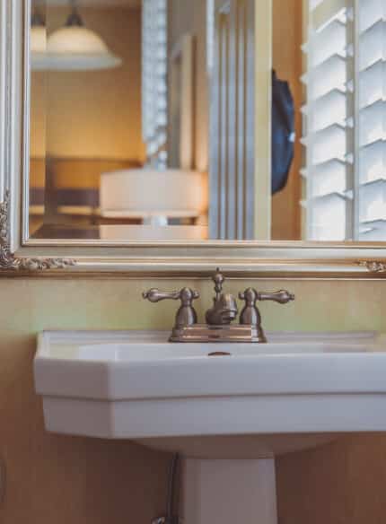 A bathroom sink with a bronze faucet is positioned below a large mirror framed in gold. There is a toilet paper holder on the left wall and white shutters on the right window.