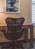 A modern desk setup with a black ergonomic chair, a wooden desk, a desk lamp, and a framed abstract painting, near two shuttered windows.