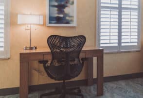 A modern desk setup with a black ergonomic chair, a wooden desk, a desk lamp, and a framed abstract painting, near two shuttered windows.