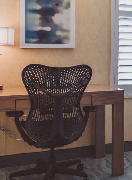 A modern desk setup with a black ergonomic chair, a wooden desk, a desk lamp, and a framed abstract painting, near two shuttered windows.