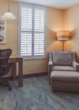 A corner room with a desk, chair, table lamp, and artwork on the wall. There's also a cushioned armchair with an ottoman and a floor lamp. The room has closed shutter windows.