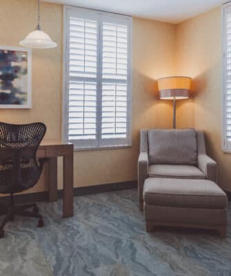 A corner room with a desk, chair, table lamp, and artwork on the wall. There's also a cushioned armchair with an ottoman and a floor lamp. The room has closed shutter windows.