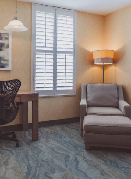 A corner room with a desk, chair, table lamp, and artwork on the wall. There's also a cushioned armchair with an ottoman and a floor lamp. The room has closed shutter windows.