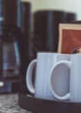 Two white coffee mugs sit on a black tray next to sealed coffee packets; a coffee maker is in the background on a marble countertop.