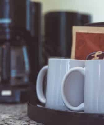 Two white coffee mugs sit on a black tray next to sealed coffee packets; a coffee maker is in the background on a marble countertop.