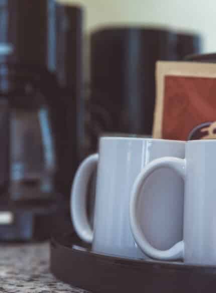 Two white coffee mugs sit on a black tray next to sealed coffee packets; a coffee maker is in the background on a marble countertop.