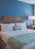 A neatly made bed with white sheets and a patterned green throw is positioned against a blue accent wall. Two side tables with lamps flank the bed, and a window with white shutters is to the right.