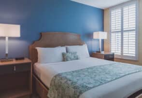 A neatly made bed with white sheets and a patterned green throw is positioned against a blue accent wall. Two side tables with lamps flank the bed, and a window with white shutters is to the right.