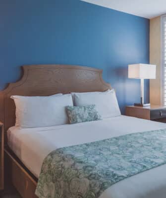 A neatly made bed with white sheets and a patterned green throw is positioned against a blue accent wall. Two side tables with lamps flank the bed, and a window with white shutters is to the right.