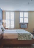 A neatly made bed with two nightstands in a well-lit room featuring light from two windows with shutters. An armchair and dresser are placed against the opposite wall.