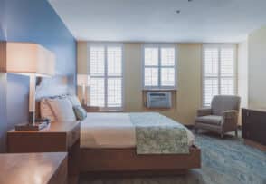 A neatly made bed with two nightstands in a well-lit room featuring light from two windows with shutters. An armchair and dresser are placed against the opposite wall.