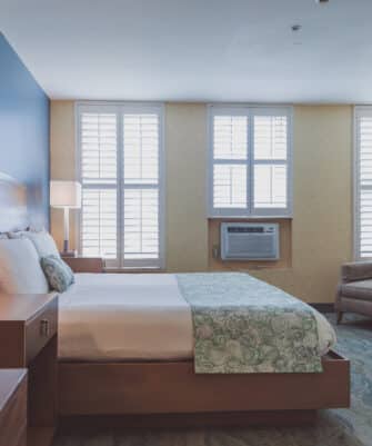 A neatly made bed with two nightstands in a well-lit room featuring light from two windows with shutters. An armchair and dresser are placed against the opposite wall.