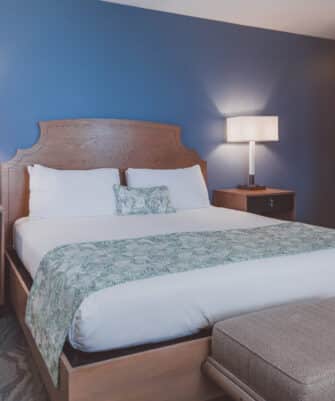 A hotel room with a large bed against a dark blue wall, flanked by two bedside tables with lamps. A cushioned bench is at the foot of the bed. The room has a modern and clean design.