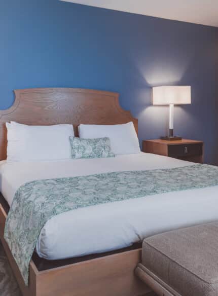 A hotel room with a large bed against a dark blue wall, flanked by two bedside tables with lamps. A cushioned bench is at the foot of the bed. The room has a modern and clean design.