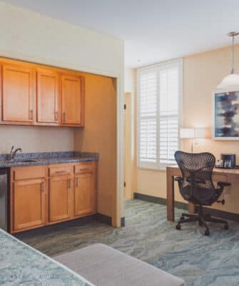 A hotel room features a small kitchenette with wooden cabinets, a microwave, and a mini fridge. A desk with a chair and lamp is situated near the window with shutters. A bed is partially visible.