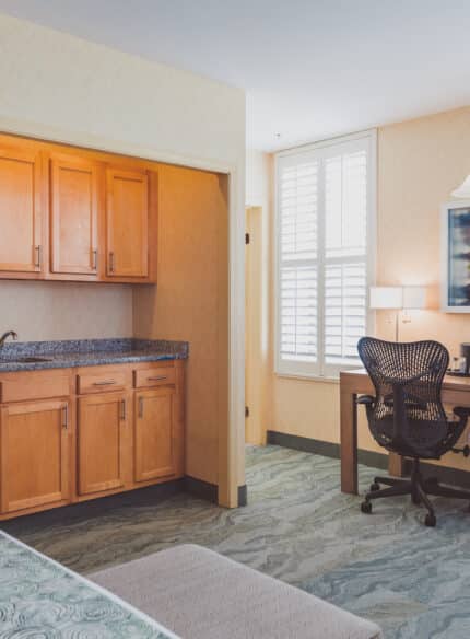 A hotel room features a small kitchenette with wooden cabinets, a microwave, and a mini fridge. A desk with a chair and lamp is situated near the window with shutters. A bed is partially visible.