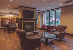 A cozy lounge area with leather chairs, a coffee table, and a fireplace in the center. Large windows let in natural light. A television is mounted above the fireplace.