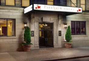 The entrance of Parkview Hotel, featuring a decorative sign, two large planters with trimmed shrubs, and flanked by two black lantern-style lights.