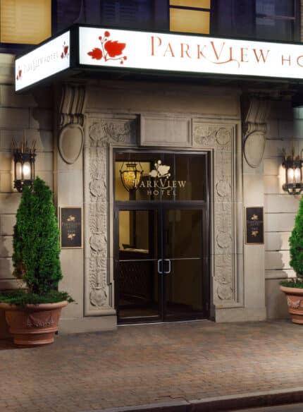 The entrance of Parkview Hotel, featuring a decorative sign, two large planters with trimmed shrubs, and flanked by two black lantern-style lights.